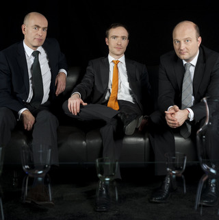 Drei Männer in schwarzen Anzügen und weißen Hemden sitzen auf einer schwarzen Ledercouch vor einem schwarzen Hintergrund.